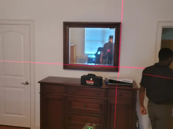 Framed mirror being hung above a dresser with red laser level lines marking the placement
