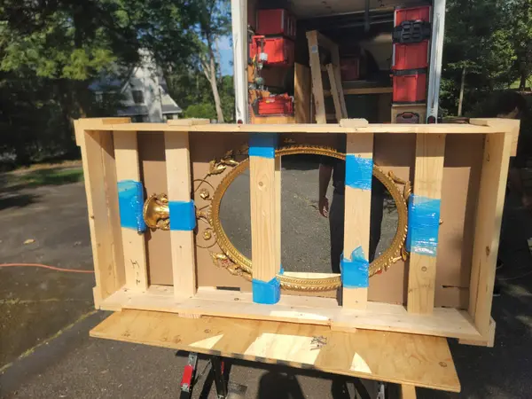 Ornate gilded mirror braced inside a custom-built wooden crate on sawhorses beside an open work truck