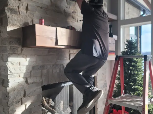 Technician on a ladder mounting a TV bracket to a stacked stone fireplace above a floating shelf